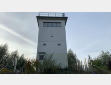 Ein alter, rechteckiger Wachturm mit flachem Dach und Geländer steht inmitten von umgebenden Bäumen und niedrigen Sträuchern. Im Vordergrund führen Treppen und Geländer zum Gebäude hoch.