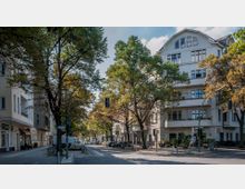 Straßenkreuzung in einem Stadtviertel mit mehrstöckigen Wohngebäuden im klassischen Stil; Bäume säumen die Straße und werfen Schatten, während geparkte Autos, Fahrräder und ein Lieferwagen sichtbar sind. Fußgänger und ein Café mit Straßenbestuhlung befinden sich im Vordergrund.