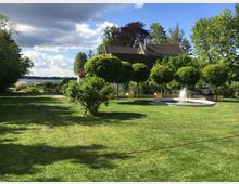 Bewohnter Garten mit gepflegtem Rasen, einem kleinen runden Springbrunnen rechts und einer Reihe von symmetrisch beschnittenen Bäumen in gelben Pflanzkübeln. Im Hintergrund ist ein Gebäude mit Schrägdach sowie ein Feld unter teilweise bewölktem Himmel zu sehen.