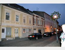 Straßenszene mit historischen Wohn- und Geschäftsgebäuden in einem Altstadtbereich, gekennzeichnet durch verzierte Fassaden mit Stuckdetails und großen Fenstern. Im Vordergrund stehen geparkte Autos entlang der gepflasterten Straße, und Straßenlaternen sind mit weihnachtlicher Dekoration geschmückt.