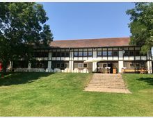 Zweistöckiges Fachwerkgebäude mit dunkelbraunem Holz und weißen Wänden, umgeben von grünem Rasen und Bäumen. Vor dem Gebäude befinden sich Treppenstufen und Tische mit Sonnenschirmen auf einer Terrasse.