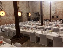 Ein großer, rustikaler Raum mit unverputzten Backsteinwänden, in dem lange, festlich gedeckte Tafeln mit weißen Tischdecken stehen. Der Raum ist mit hölzernen Säulen, schlichten Kronleuchtern und Blumendekorationen geschmückt, was auf eine Veranstaltung wie eine Hochzeit oder ein Bankett hindeutet.