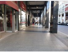 Überdachter Gehweg mit großen, dunklen Säulen entlang einer Einkaufsstraße. Rechts parken Autos am Straßenrand, links befinden sich Schaufenster und ein Restaurant mit roter Markise.