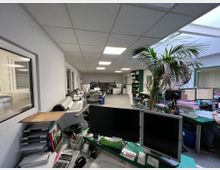 Großraumbüro mit mehreren Arbeitsplätzen, Computermonitoren und einem großen Drucker an der linken Wand. Der Raum ist hell beleuchtet durch Deckenlampen und ein Oberlicht; im Hintergrund befinden sich Regale mit Aktenordnern und weitere Arbeitsbereiche.