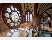 Innenraum einer umgebauten Kirche mit roten Ziegelwänden, einem großen runden Rosettenfenster und gotischen Spitzbogenfenstern. Der Raum verfügt über eine moderne Treppenkonstruktion aus Holz und Metall sowie eine Orgel im Hintergrund, die den historischen und modernen Charakter des Gebäudes kombiniert.