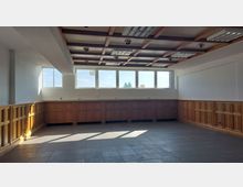 Leerer Innenraum mit großen rechteckigen Fenstern, dunklen Bodenfliesen und einer Holzvertäfelung an den Wänden. Die Decke ist mit sichtbaren Holzbalken und eingelassenen Leuchten gestaltet.