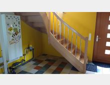 Treppenhaus mit gelb gestrichenen Wänden, einer Holztreppe mit Metallgeländer und einem Boden aus mehrfarbigen quadratischen Fliesen. Links steht ein Kühlschrank mit Magneten, rechts eine Holztür mit eingelassenem Fensterdesign.