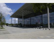 Ein modernes Gebäude mit einer großen Glasfassade und einem weit auskragenden Flachdach, getragen von schmalen Säulen. Der Vorplatz ist mit Betonplatten und Pflastersteinen gestaltet, umgeben von vereinzelten Bäumen und Sitzgelegenheiten.