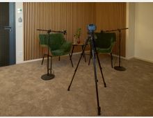 Ein kleiner Aufnahmeraum mit grauem Teppichboden, an einer Wand mit Holzpaneelen. Zwei grüne Sessel um einen kleinen Tisch mit Pflanzen und Wasserflasche, ausgestattete Mikrofone und ein Stativ mit Kamera in der Mitte.