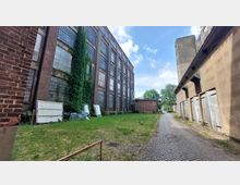 Industriegelände mit einem alten Backsteingebäude auf der linken Seite, dessen Fassade teilweise mit Efeu bewachsen ist, und einem weiteren Gebäude mit verblassten Putz- und Holztüren auf der rechten Seite. Dazwischen verläuft ein gepflasterter Weg, daneben eine Rasenfläche mit vereinzeltem Bewuchs.