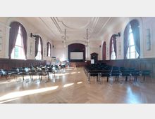 Ein historisch wirkender Veranstaltungsraum mit hohen Decken, holzgetäfelten Wänden und großen Fenstern mit burgunderfarbenen Vorhängen. Reihen von schwarzen Stühlen sind auf einen Bühnenbereich mit Projektionsleinwand, roten Vorhängen und mehreren Werbebannern ausgerichtet.