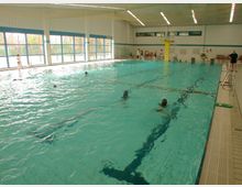 Halle eines Hallenbads mit einem großen Schwimmbecken, Markierungen für Schwimmbahnen, Sprungturm im Hintergrund und großen Fenstern, die Tageslicht hereinlassen. Mehrere Personen schwimmen oder stehen am Beckenrand.