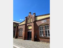 Backsteinbau mit gelber Fassadenakzentuierung und spitzem Ziergiebel, großen Fenstern mit Gitter und einer offenen Eingangstür. Der Vorplatz ist mit Kopfsteinpflaster gestaltet, und an der Wand befindet sich eine Hausnummer (5) sowie ein Schild mit Informationen.