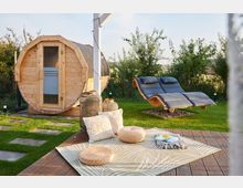 Holzfasssauna in einem Garten mit Rasenfläche, daneben eine geschwungene Outdoor-Liege mit Polsterung. Im Vordergrund liegt ein mit Kissen und runden Sitzkissen dekorierter Outdoor-Teppich auf einer Holzterrasse.