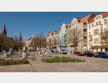 Offener Platz mit Kopfsteinpflaster, umgeben von mehrstöckigen Wohnhäusern mit farbigen Fassaden und roten Dächern; auf dem Platz stehen Bäume, Kunstinstallationen aus Stein und eine Straßenlaterne. Im Hintergrund sind die Türme einer Kirche zu sehen, eingebettet in Bäume und blauen Himmel.