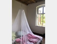 Ein einfacher Raum mit Holzboden und einer unverputzten Holzdecke. Im Vordergrund steht ein Bett mit buntem Bettzeug und einem weißen Moskitonetz, daneben ein geöffnetes Fenster mit Blick ins Grüne.