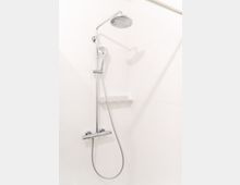 Moderne Dusche mit verchromtem Duschkopf und Handbrause an einer weißen, gefliesten Badezimmerwand. Darunter befindet sich eine Mischarmatur, und ein kleines Regal ist an der Wand angebracht.