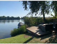 Holzterrasse mit Gartenmöbeln am Ufer eines ruhigen Sees, umgeben von Gras, Sträuchern und einem Baum. Im Hintergrund sind Bäume und das gegenüberliegende Ufer sichtbar, ein kleines Boot mit Plane steht am Ufer.