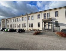 Zweistöckiges weißes Gebäude mit großen, rechteckigen Fenstern und einem Vordach über dem Haupteingang, der über eine Treppe erreichbar ist. Im Vordergrund befindet sich ein Parkplatz mit mehreren Autos und angrenzenden niedrigen Hecken.