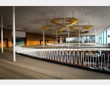 Geräumiger Innenbereich eines modernen Gebäudes mit einer geschwungenen Empore, die von weißen Geländerstreben umgeben ist. Die Decke ist mit großen, dekorativen, goldfarbenen Leuchten gestaltet, und große Fenster auf der rechten Seite lassen viel Tageslicht herein.