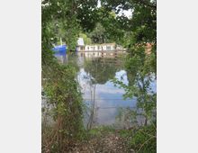 Blick durch dichte Bäume und Sträucher auf einen ruhigen Fluss mit einem blauen Boot und einem Hausboot im Hintergrund. Die Umgebung ist grün und spiegelt sich im Wasser.