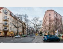 Straßenszene in einer städtischen Umgebung mit mehrstöckigen historischen Wohnhäusern auf beiden Seiten, einigen Geschäften im Erdgeschoss und geparkten Autos entlang der Straße. Bäume ohne Laub säumen die Straße, und der Gehweg ist gepflastert.