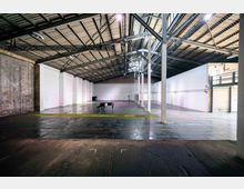 Innenansicht einer großen, leeren Industriehalle mit hohen Decken und sichtbaren Stahlträgern. Die Wände sind aus weißem Putz und Backstein, der Boden reflektiert Licht, und ein einzelnes Klavier steht im hinteren Bereich der Halle.