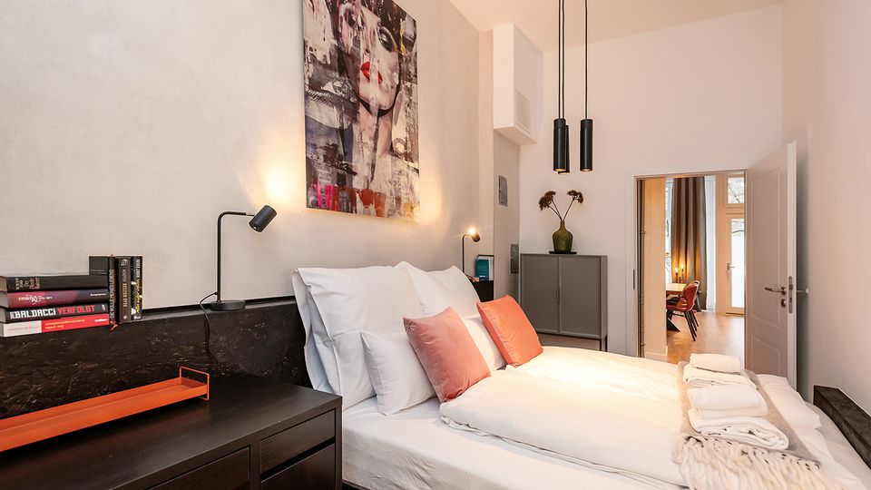 Modernes Schlafzimmer mit Doppelbett, weißen Wänden und dunklen Holzschränken; über dem Bett hängt ein großes Wandbild, daneben stehen Nachttischlampen. Zwei schwarze Pendelleuchten hängen von der Decke, und eine offene Tür führt in einen angrenzenden, hellen Raum mit Holzboden.