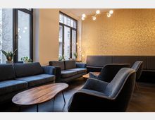 Ein modern eingerichteter Lounge-Bereich mit dunkelblauen Polstermöbeln, Holztischen und großen Fenstern, durch die Tageslicht einfällt. Die Wand ist mit goldener Tapete gestaltet, und Zimmerpflanzen sowie eine minimalistische Lampe ergänzen das Ambiente.