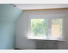 Ein leerer Raum mit einer großen rechteckigen Fensterfront, durch die grüne Bäume sichtbar sind. Unter dem Fenster befindet sich ein Heizkörper, die Wände sind teils hellblau und teils beige tapeziert.