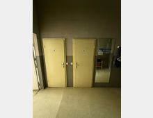 Innenraum eines Flurs mit zwei nebeneinanderliegenden, hellgelben Toilettentüren mit Piktogrammen an einer grauen Wand. Rechts hängt ein großer Wandspiegel neben einer Garderobe; der Boden ist hell und glatt.