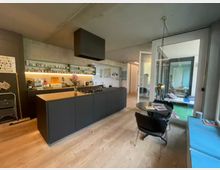 Moderne Wohnküche mit schwarzer Kücheninsel, Holzarbeitsplatte und eingelassenem Gasherd. Im Hintergrund Regale mit Geschirr, ein Esstisch mit Stühlen und eine verglaste Tür zu einem weiteren Wohnbereich.