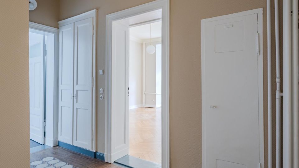 Heller Wohnungsflur mit beigefarbenen Wänden, weiß gerahmten Türen und Einbauschrank sowie sichtbaren Rohrleitungen an der rechten Wand. Durch eine offene Tür blickt man in einen weiteren Raum mit Parkettboden und Deckenleuchte.