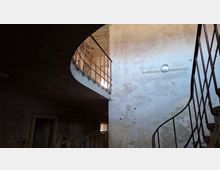 Innenansicht eines heruntergekommenen Treppenhauses in einem alten Gebäude mit abblätterndem Putz und rissigen Wänden. Rechts führt eine geschwungene Metalltreppe nach oben zu einem halb offenen Rundgang mit Geländer, beleuchtet durch ein kleines Fenster und eine einfache Wandlampe.