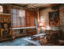 Ein gemütlicher Raum im Vintage-Stil mit rauer Backsteinwand und einem großen Fenster mit Holzjalousien. Das Interieur umfasst Ledersessel, Holzstühle, einen Teppich mit geometrischem Muster, eine Stehleuchte und dekorative Wandkunst, darunter ein Retro-Poster und eine bunte Wandteppichszene.