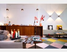 Ein gemütlicher Innenraum mit dekorativen Elementen: Im Vordergrund ein Tisch mit Zeitschriften und brennenden Kerzen in verschiedenen Farben, daneben zusammengelegte Decken. Im Hintergrund ein Sideboard aus Holz mit Dekorationen wie roten Zweigen in einer Vase und einer Weinflasche. An der Wand hängen drei Leuchten, darunter Sitzkissen mit Comic-Motiven. Der Boden ist mit farbigen, geometrischen Fliesen gestaltet.