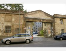 Backstein-Gebäudeeingang mit einem großen, verwitterten Torbogen, flankiert von zwei kleinen Fenstern mit Gittern; das Tor führt zu einem gepflasterten Innenhof mit weiteren Gebäuden im Hintergrund. Im Vordergrund stehen zwei geparkte Autos an einer Straße.