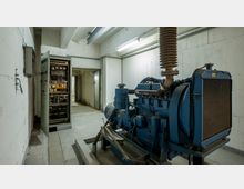 Ein technischer Raum mit Betonwänden, einer großen blauen Maschine (Generator) rechts im Vordergrund und einem offenen Schaltschrank mit elektrischem Equipment auf der linken Seite. Im Hintergrund führt eine offene Tür in einen weiteren Betonraum.