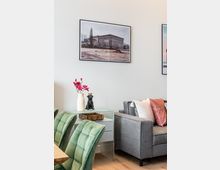 Heller Wohn- und Essbereich mit grauem Sofa und farbigen Kissen, daneben ein kleiner Beistelltisch mit Vase und Dekoobjekt. Im Vordergrund stehen grüne Polsterstühle am Holztisch; an der weißen Wand hängen gerahmte Fotografien.