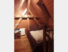 Ein Schlafbereich in einem Dachzimmer mit holzverkleideten Wänden und Decken, sichtbaren Holzbalken und einem schrägen Dachfenster. Das Zimmer enthält ein Bett mit heller Bettdecke, ein kleiner Tisch mit gefalteter Wäsche und einen Heizkörper.
