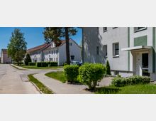 Wohnsiedlung mit mehrstöckigen, hell verputzten Wohnhäusern entlang einer gebogenen Gehweg- und Straßenführung. Vor dem rechten Gebäude sind ein Eingang mit Vordach, Grünstreifen, Hecken und einzelne Bäume zu sehen.