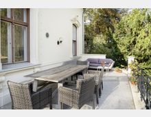 Terrasse mit cremefarbener Hausfassade, einem großen Holztisch und sechs geflochtenen Stühlen. Im Hintergrund befindet sich eine Sitzgruppe mit Sofa und Beistelltisch, umgeben von üppiger Vegetation und einem schmiedeeisernen Geländer.
