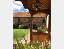 Ein Innenhof mit einem gepflasterten Weg und einer Rasenfläche, umgeben von einer Backsteinfassade mit grünen Fensterläden. Im Vordergrund befindet sich eine Holzüberdachung mit einem hängenden Wagenrad und dekorativen Elementen, darunter eine alte Waage und Pflanzen.