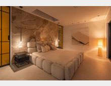 Modernes Schlafzimmer mit großem gepolstertem Bett und heller Bettwäsche vor einer großformatigen Marmor-Akzentwand. Warmes indirektes Licht, schmale Nachttische, gelb getönte Glas-Elemente und ein großes abstraktes Wandbild sowie eine Stehlampe prägen den minimalistischen Raum.