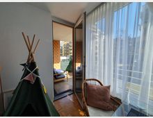 Ein heller Raum mit bodentiefen Fenstern und transparenten Vorhängen, der auf einen Balkon mit Sitzmöbeln und einem Sichtschutz führt. Im Vordergrund stehen ein kleines grünes Spielzelt und ein Rattanstuhl mit einem braunen Kissen.
