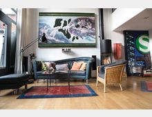 Wohnzimmer mit hellen Holzböden und modernen Elementen, darunter ein dunkelblaues Ledersofa, ein Rattansessel und ein Glastisch. An der Wand hängt ein modernes Kunstwerk, daneben befinden sich ein Kamin mit Holzvorrat und eine große Fensterfront, die viel Licht bietet.