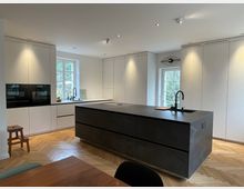 Moderne Küche mit hellen, glatten Einbauschränken und großen Fenstern, die Tageslicht hereinlassen. Im Zentrum steht eine dunkle Kücheninsel mit integriertem Waschbecken, flankiert von einem Fischgrät-Holzboden.