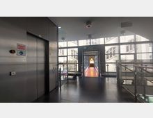 Ein moderner Innenraum mit einem Fahrstuhl auf der linken Seite, großen Fensterfronten im Hintergrund und einem überdachten Übergang mit rotem Boden, der zu einem angrenzenden Gebäude führt. Der Bereich ist mit dunklen Fliesen und Geländern aus Metall ausgestattet.