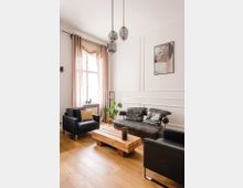 Ein modernes Wohnzimmer mit Holzboden, schwarzen Ledersesseln und einem passenden Sofa. Ein rechteckiger Couchtisch aus Holz steht in der Mitte, darüber hängen drei stilvolle Glaskugelleuchten, und im Hintergrund ist ein großes Fenster mit cremefarbenen Vorhängen.