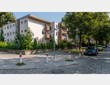 Eckansicht eines hellen Wohngebäudes mit Flachdach, mehreren Balkonen und kleinen Fenstern, umgeben von Hecken und Bäumen. Vor dem Gebäude liegt eine asphaltierte Straße mit Bordsteinen, Gehwegpollern und parkenden Autos entlang einer von Bäumen gesäumten Straße.